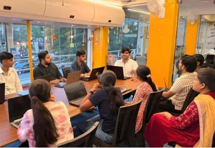 People sit around a conference table with laptops in a brightly lit office.